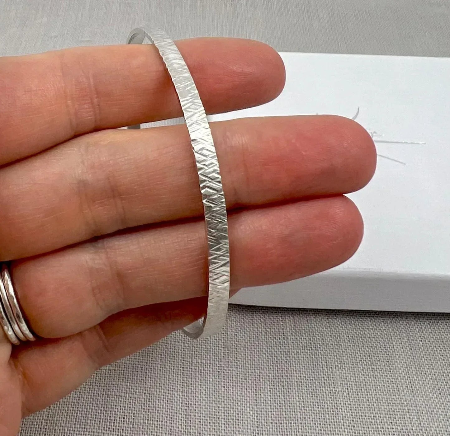 Hand holding a textured silver criss-cross bangle bracelet on light fabric background