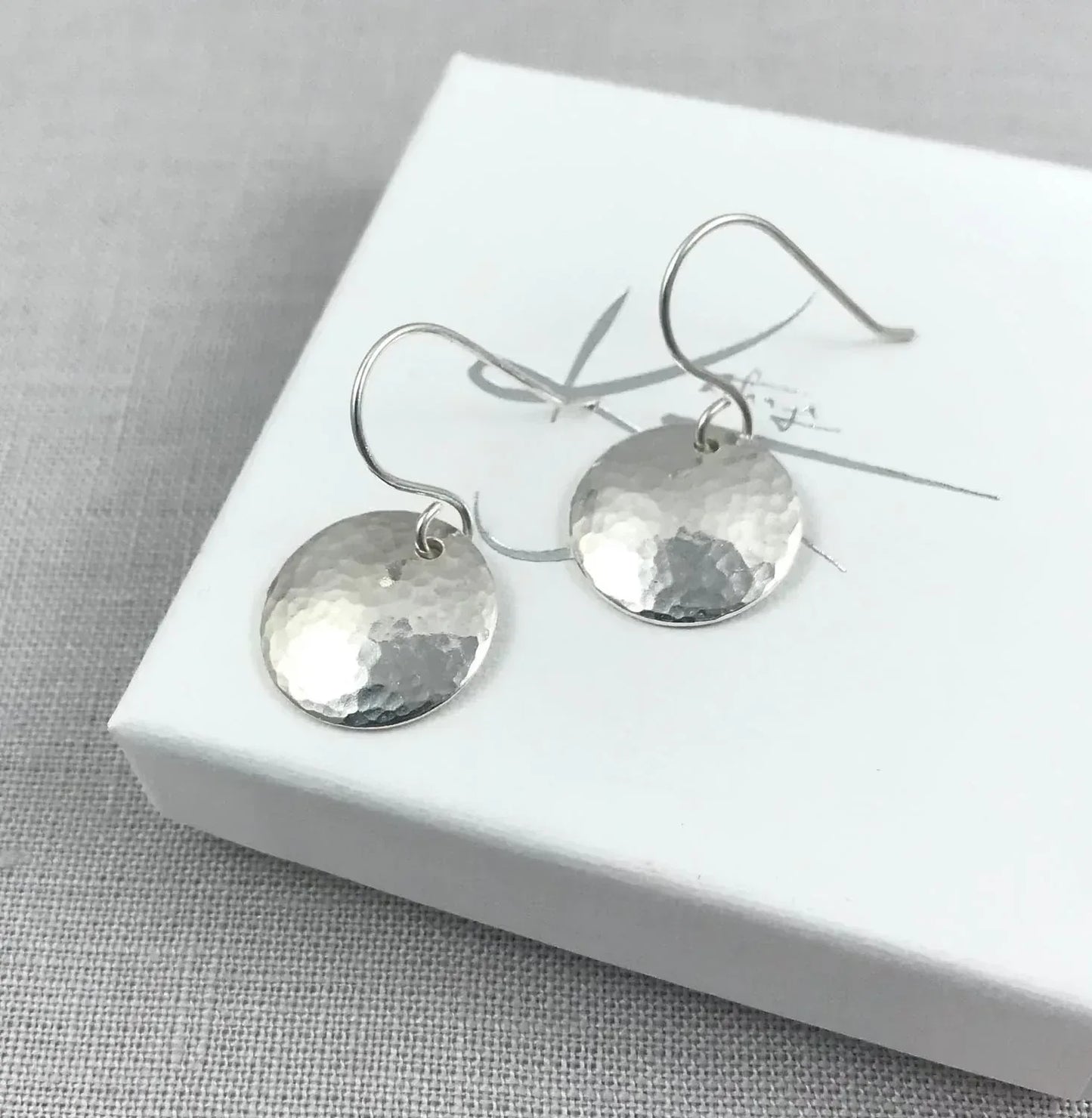 Hammered silver dome drop earrings on white jewelry box with textured background