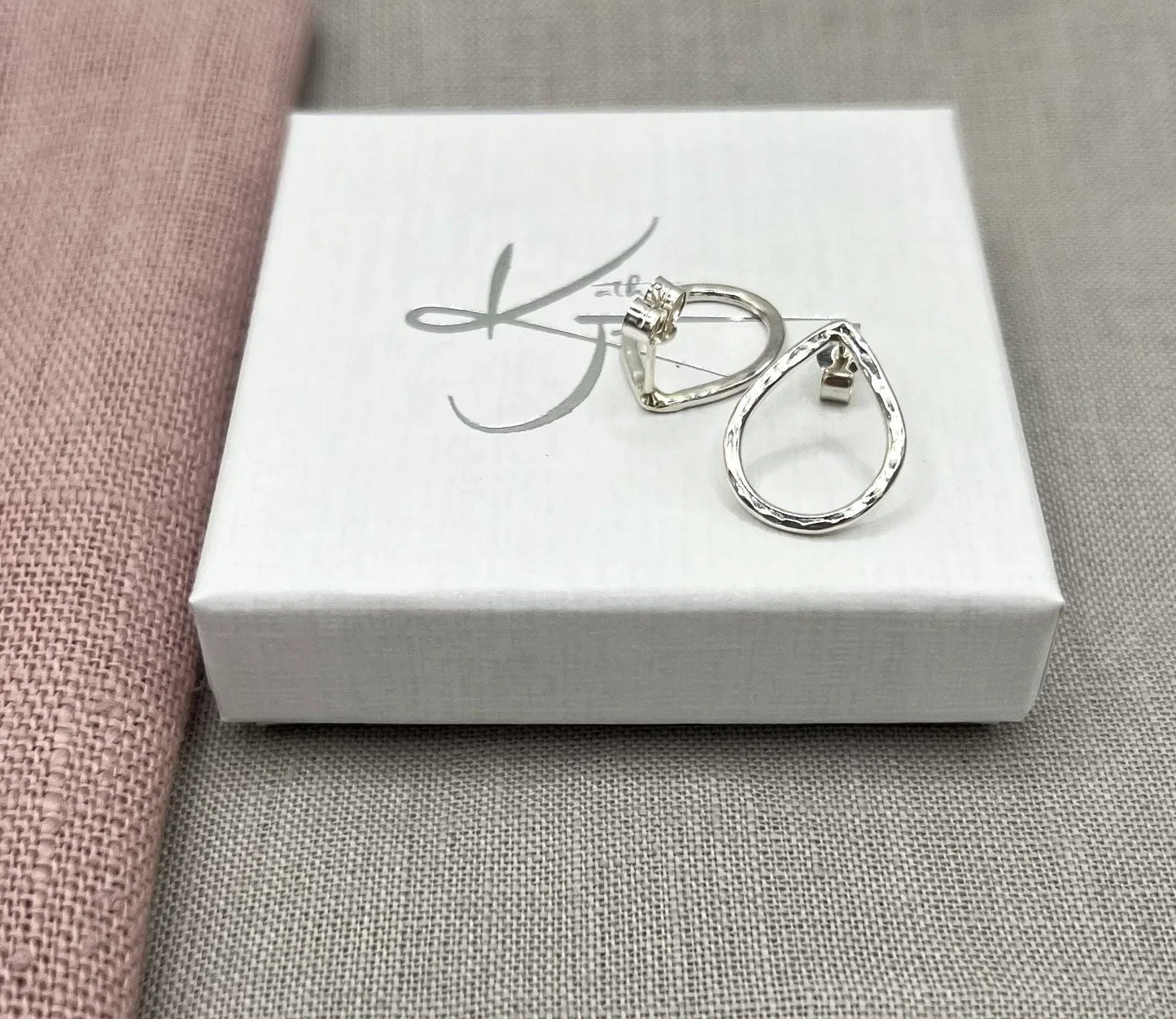 Hammered silver teardrop stud earrings on a white jewelry box with pink and grey fabric background