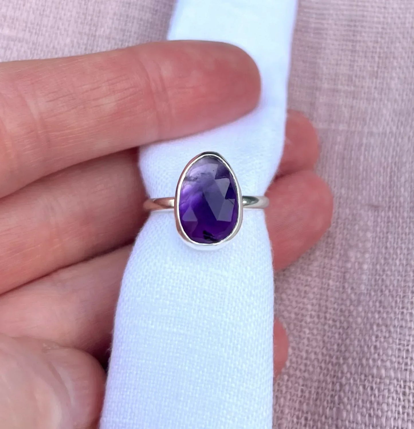 Hand holding silver ring with oval faceted purple gemstone on white fabric