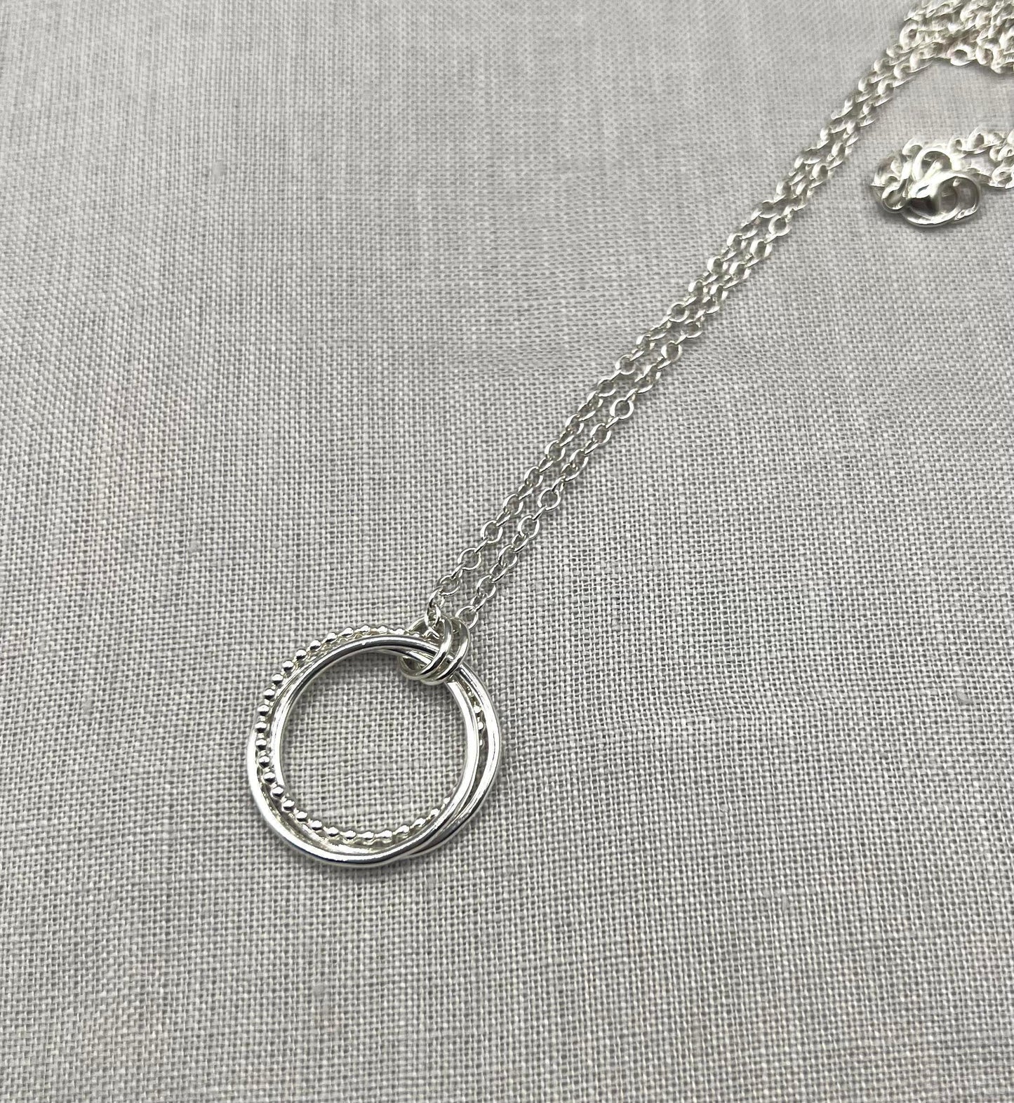 Interlinked silver hoop necklace with textured rings on a fine chain, displayed on linen.