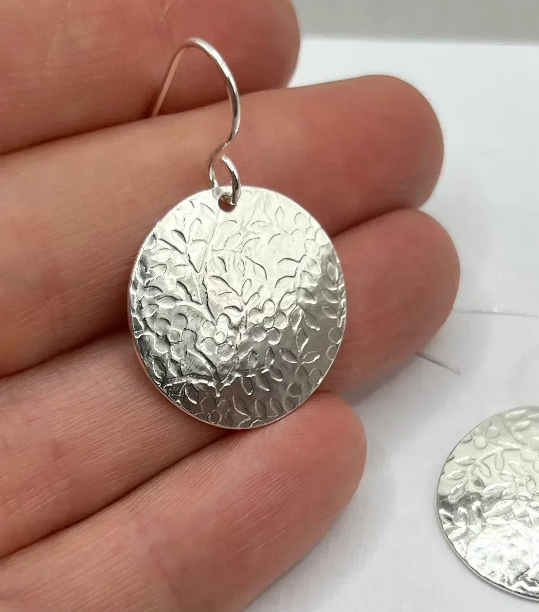 Hand holding a silver disc earring with embossed floral pattern
