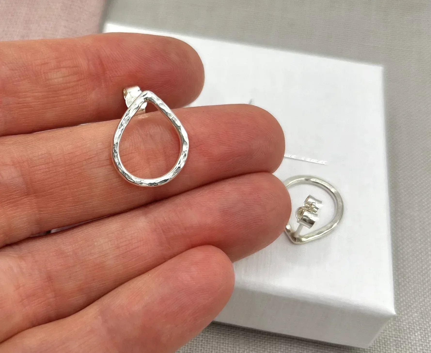 Hand holding a hammered silver teardrop stud earring with another displayed on a box