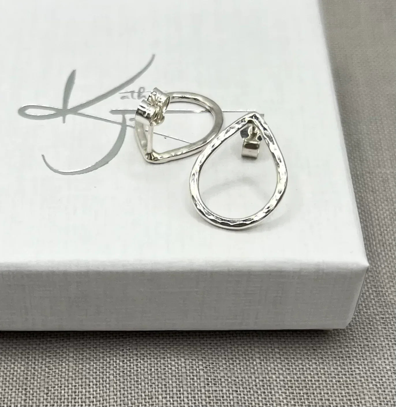 Hammered silver teardrop stud earrings on a white jewelry box, textured finish