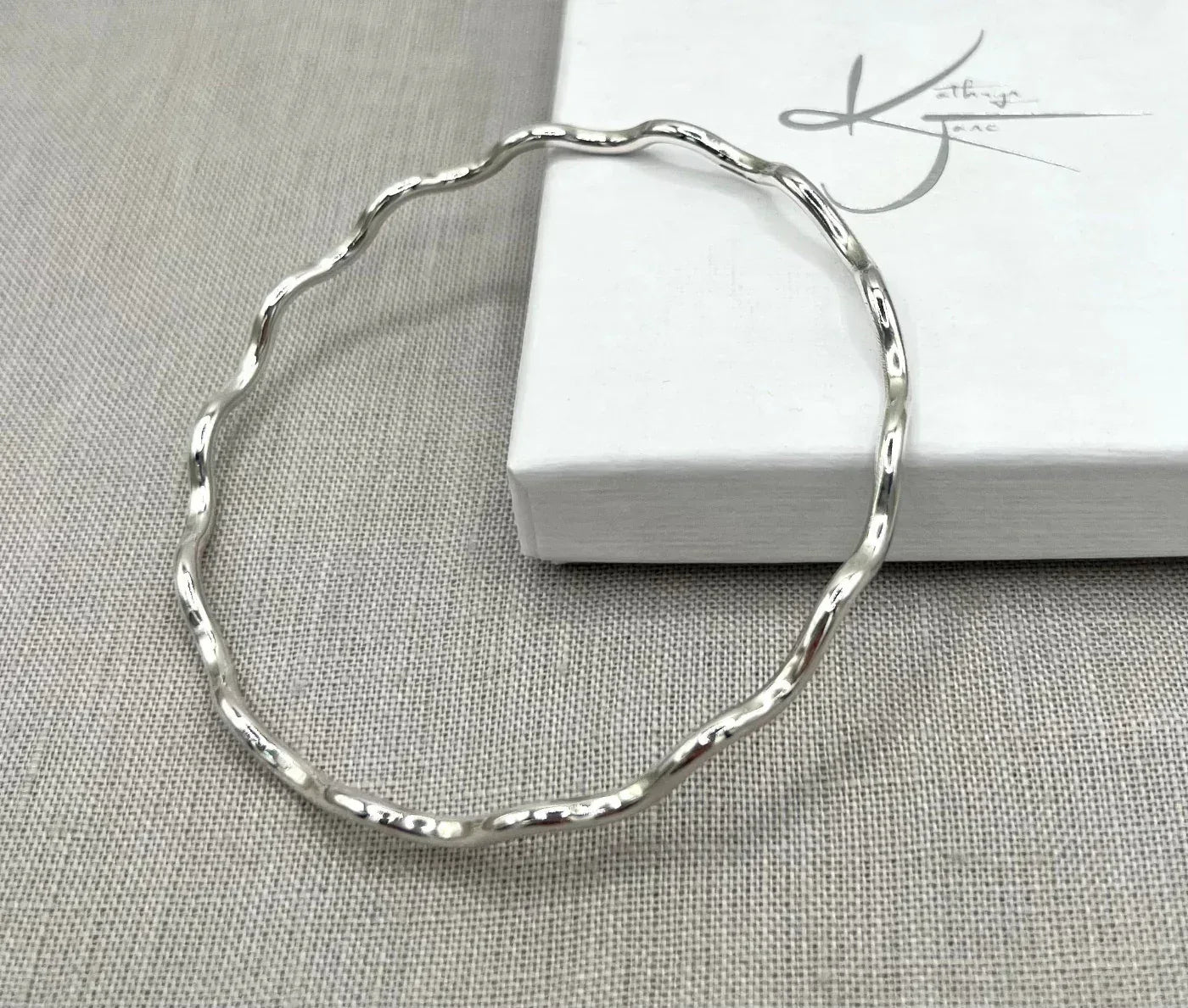 Sterling silver wavy bangle bracelet on light fabric beside a white jewelry box