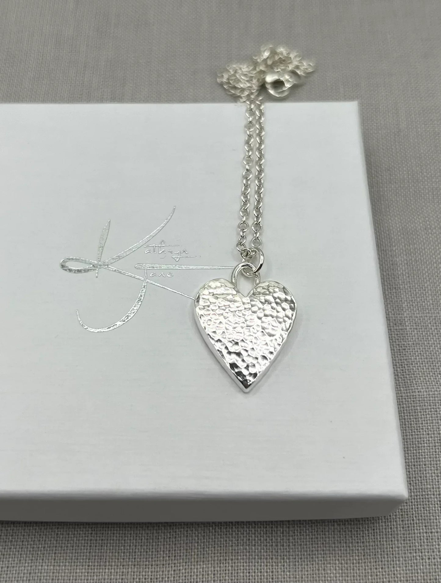 Hammered silver heart pendant necklace on chain, displayed on white jewelry box.