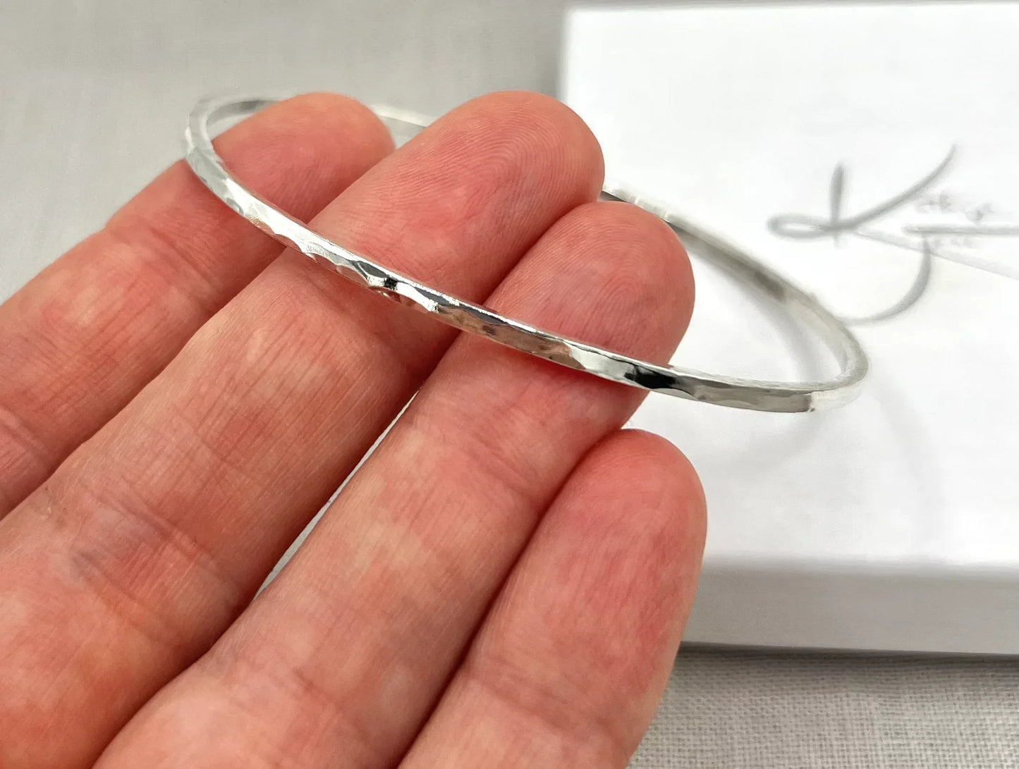 Hand holding a lightly hammered silver square wire bangle in front of a white box