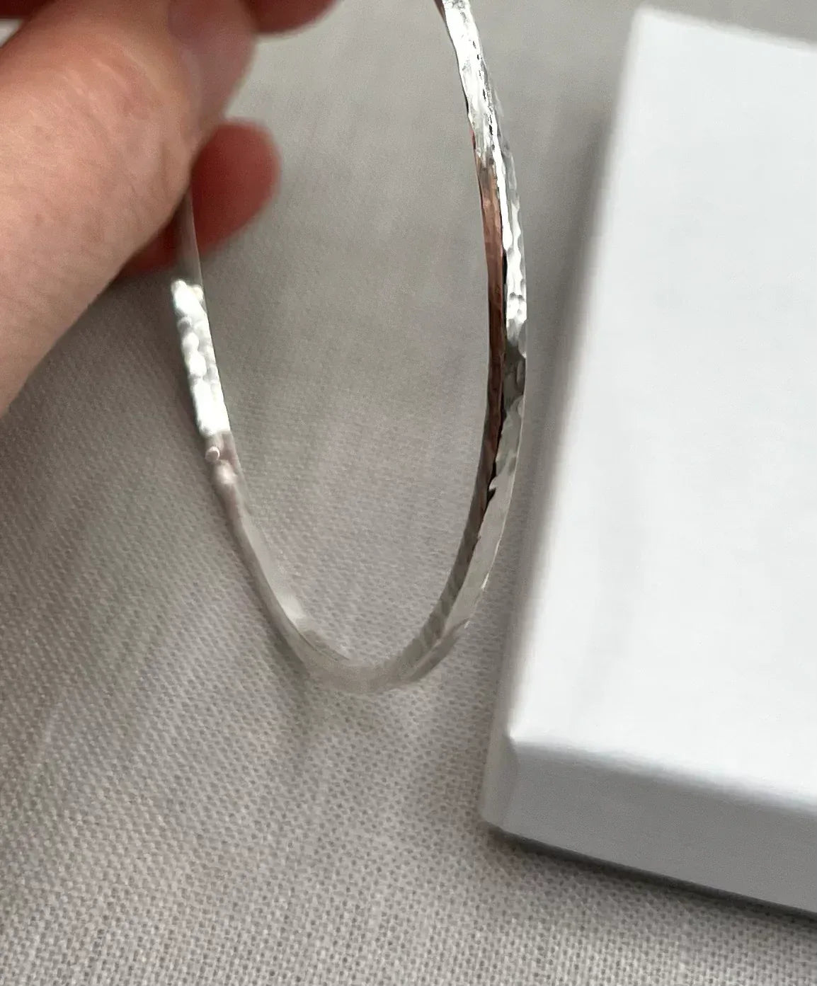 Hand holding a hammered silver twist bangle on linen with a white gift box
